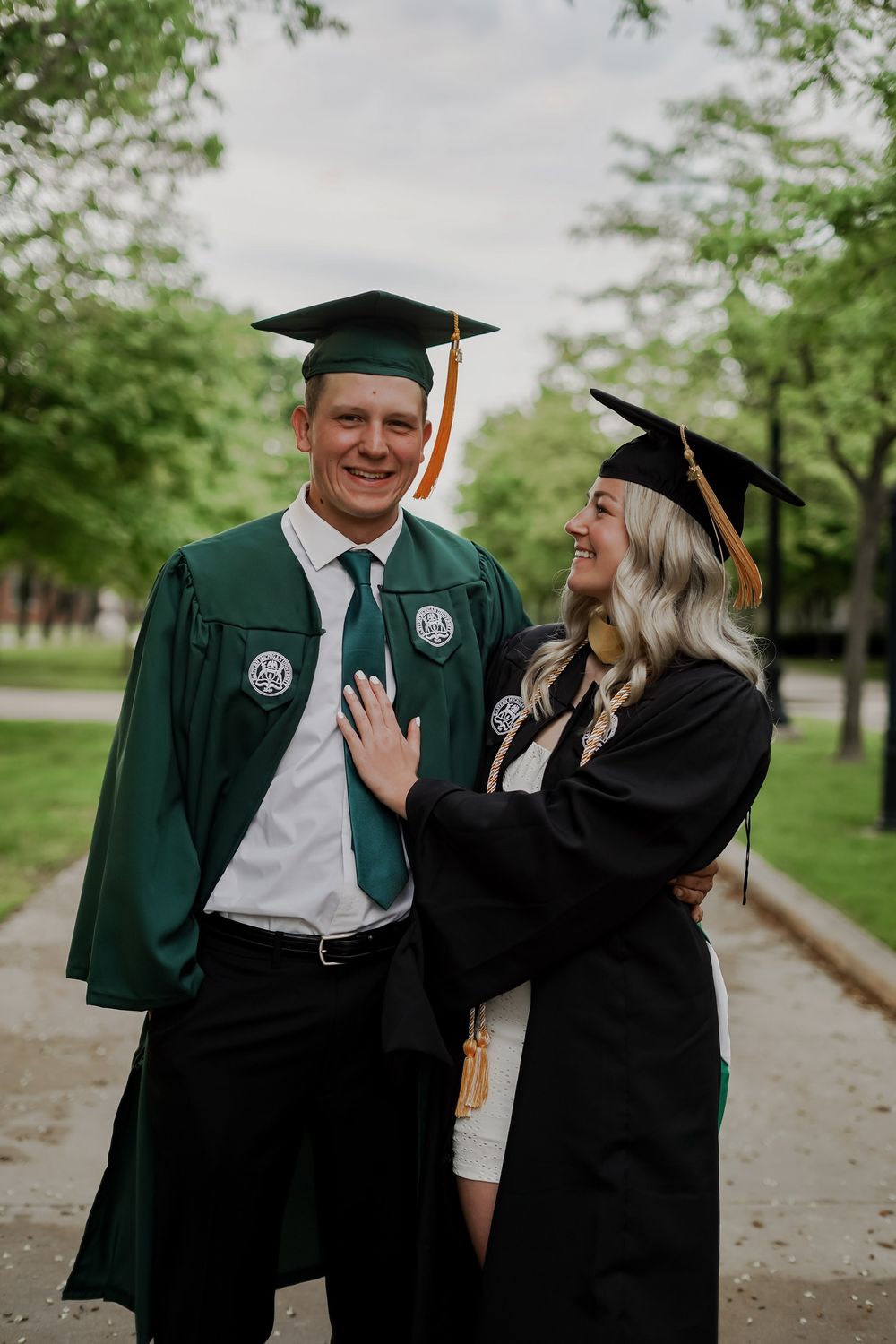 Graduates in green and black commencement robes share joyful moment outdoors.