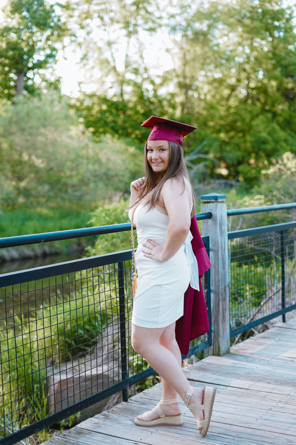 Graduate in burgundy regalia sits on wooden boardwalk surrounded by greenery.