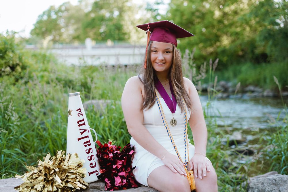 Graduate in burgundy cap and gown poses outdoors with floral bouquet against natural backdrop.