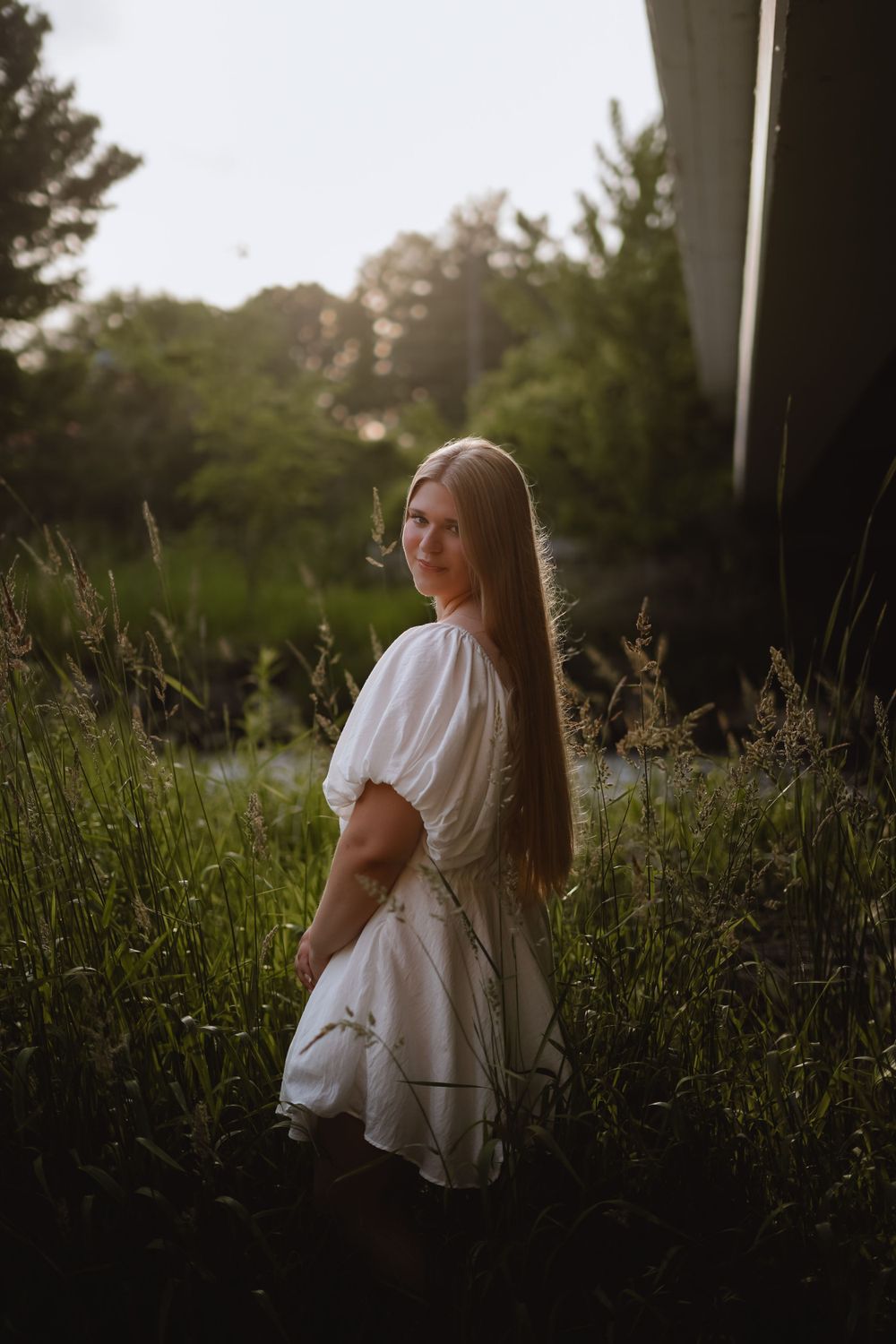 Person in white dress sitting in tall grass during sunset photoshoot.