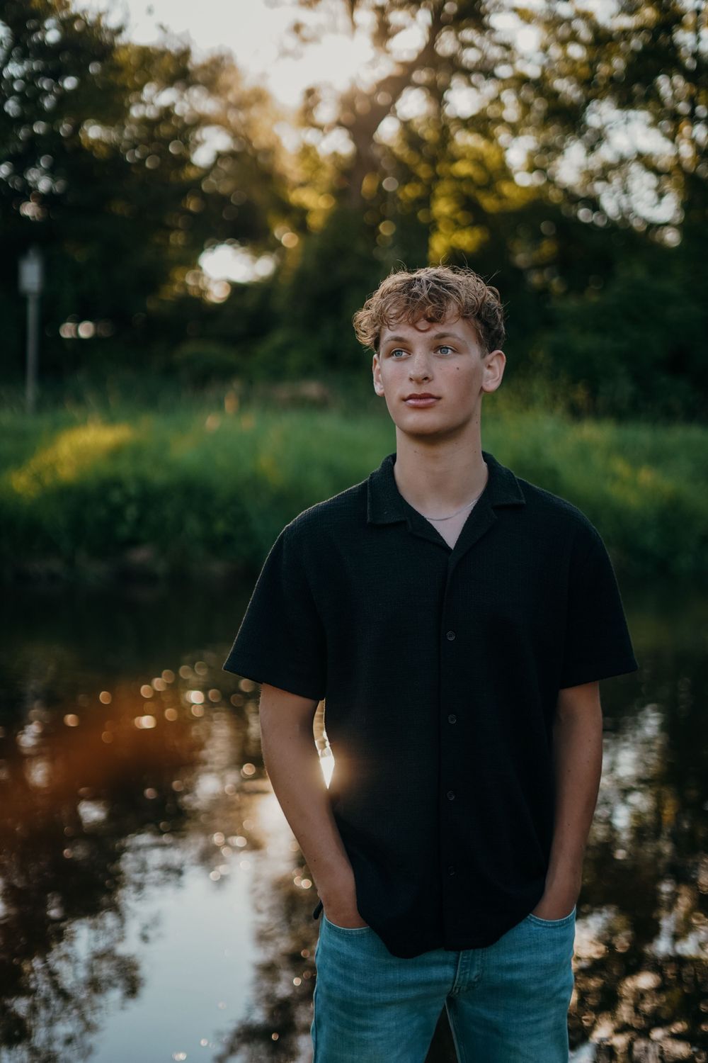 Person in black polo and jeans posing by water during golden hour.