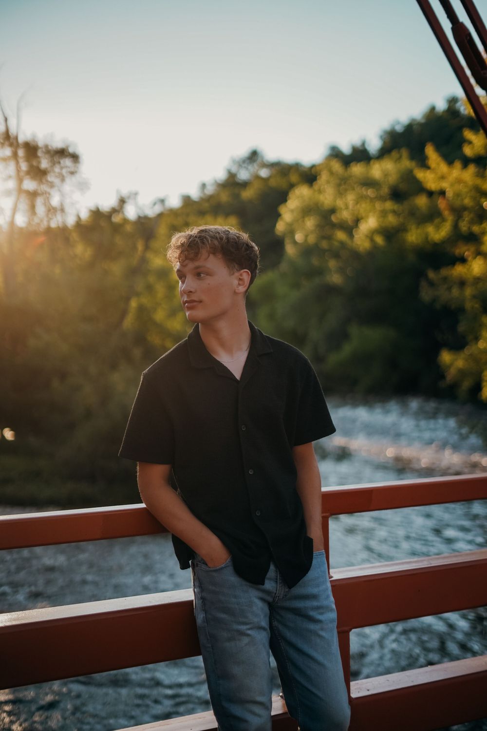 Person in black polo shirt leaning against railing by water at sunset.