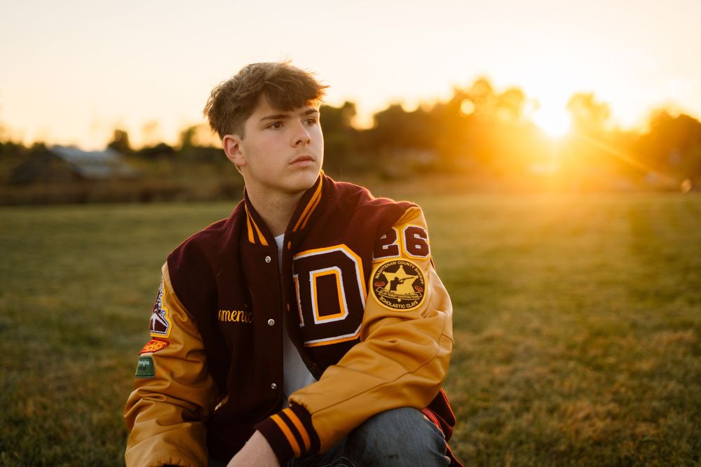 Person wearing letterman jacket poses in golden sunlight outdoors.