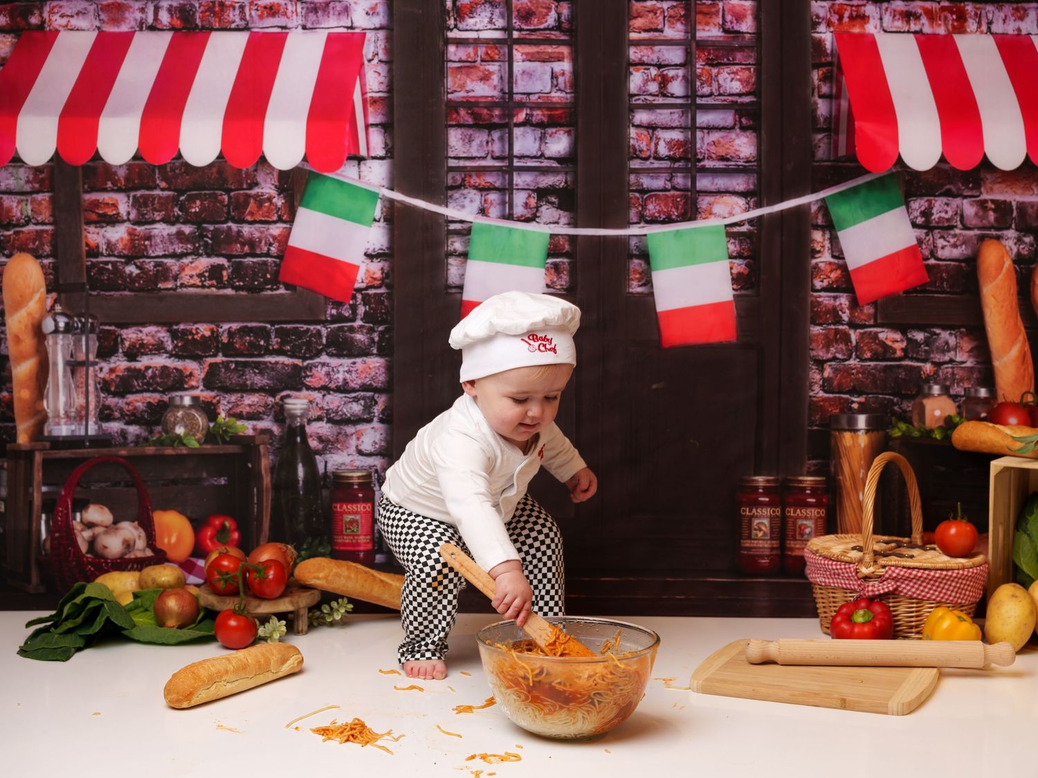 A small child in chef attire stirs a bowl while Italian flags hang above in a rustic kitchen setting.