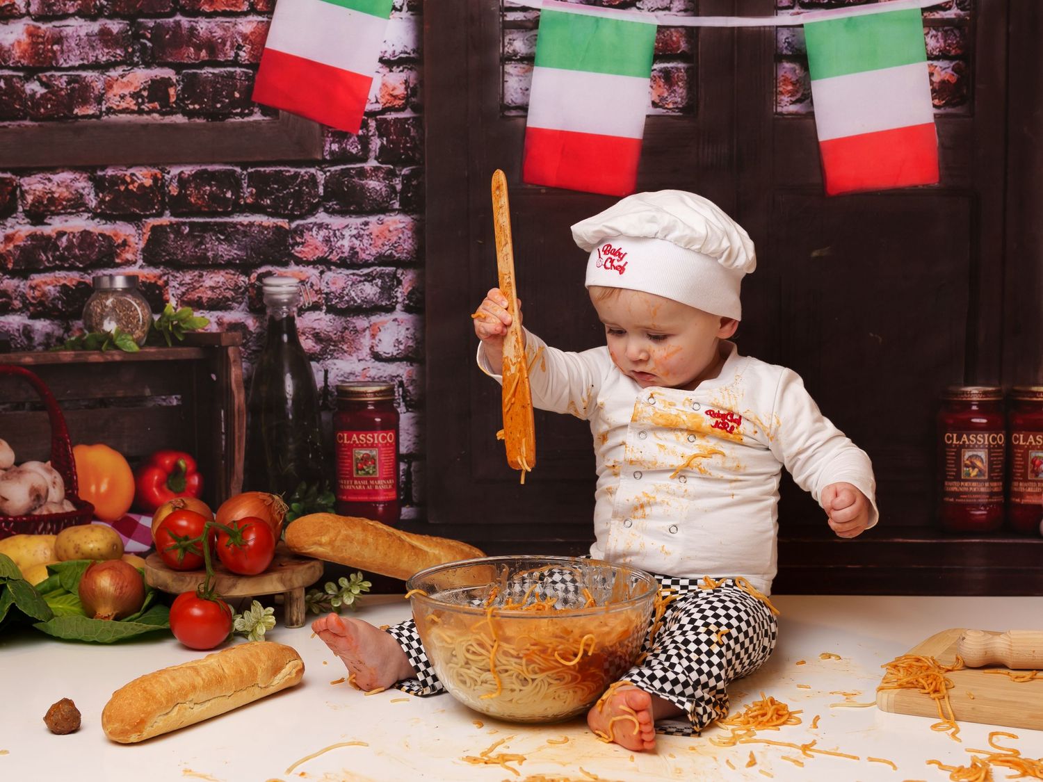 A toddler in a chef hat holds a wooden spoon while sitting on a counter with Italian flags and ingredients.