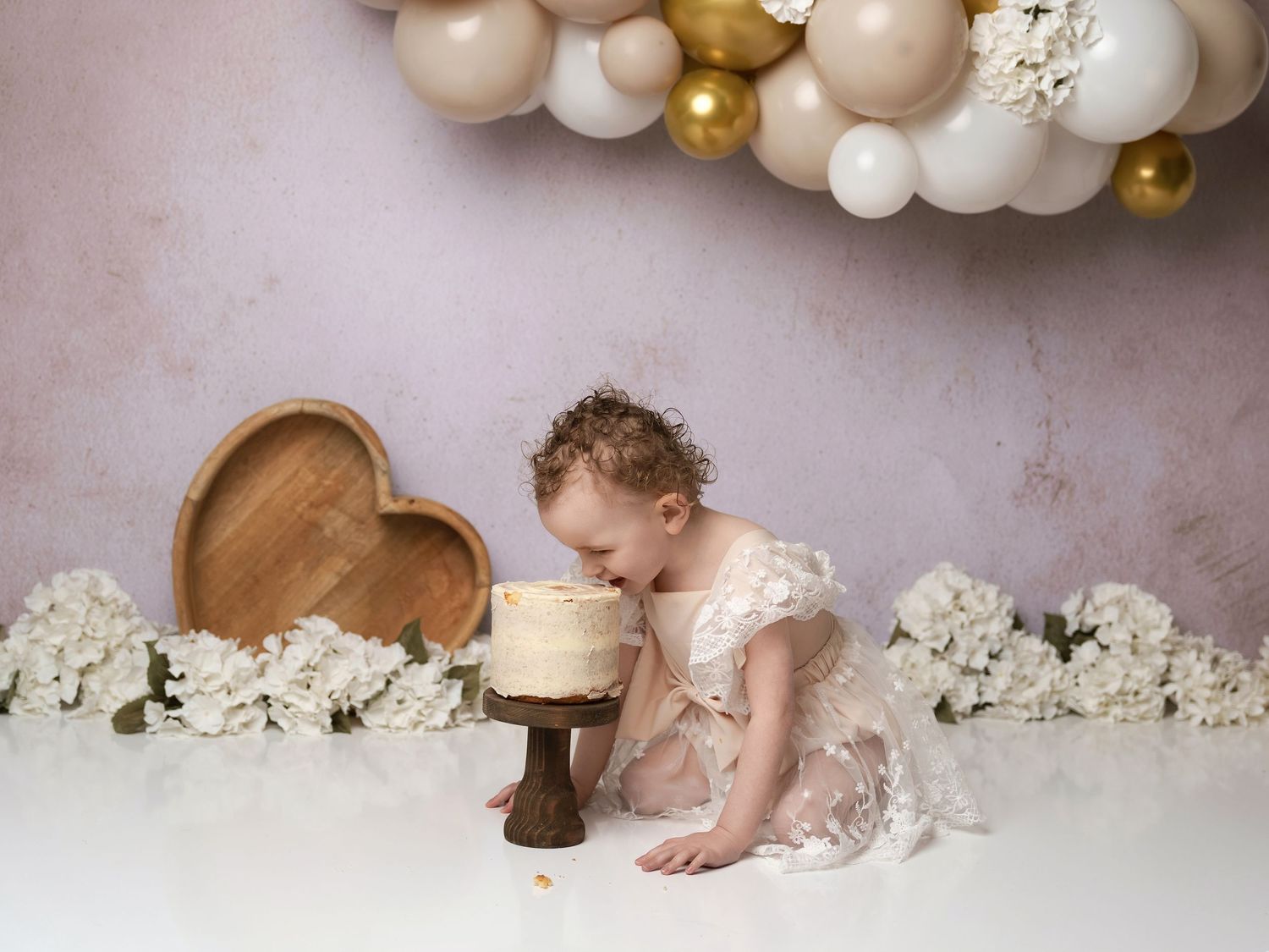 Whimsical first birthday setup with balloon arch, wooden heart, white florals and cake on pedestal against pink wall.