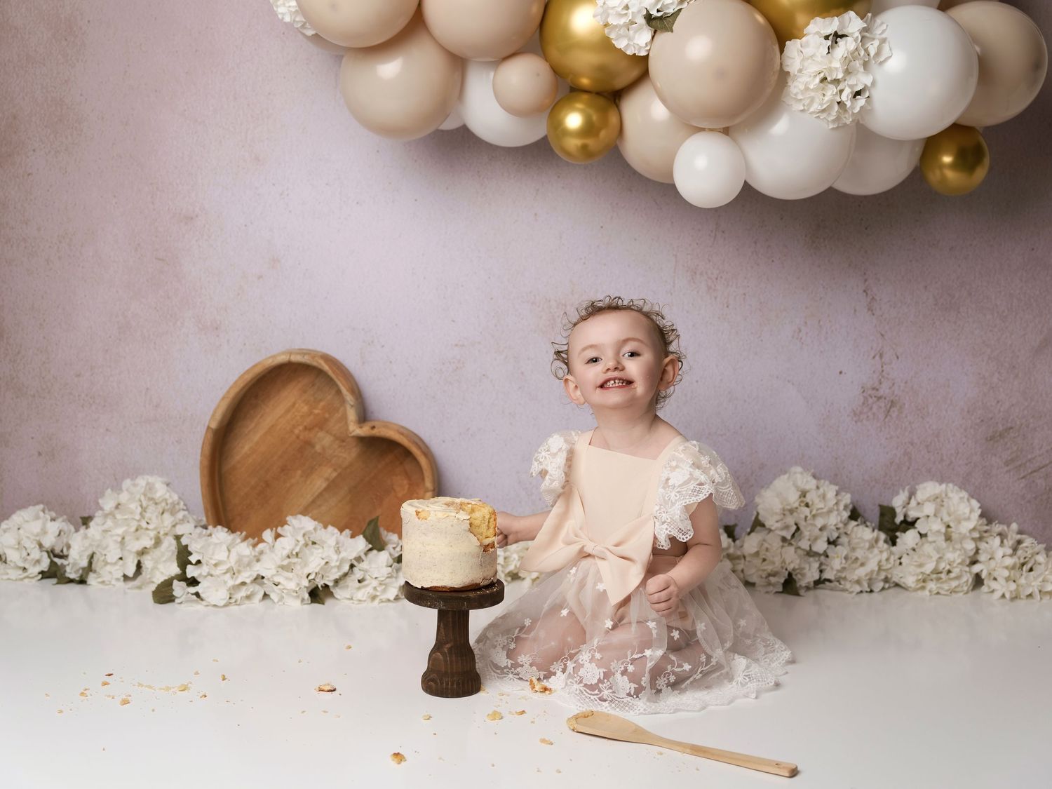 A cake smash photo setup with white and gold balloon arch, floral decor, and wooden accents on a neutral backdrop.