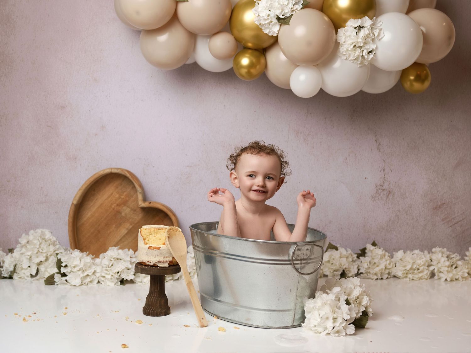 Beautiful cake smash setup with gold and white balloon arch, flowers, rustic decor and metallic washtub against neutral backdrop.