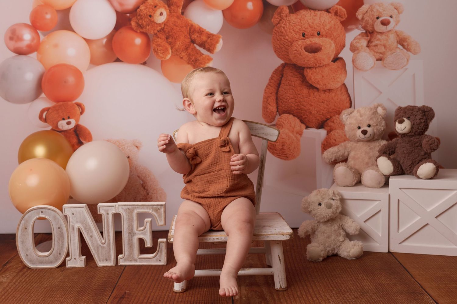 First birthday setup with orange and white balloons, teddy bears, and wooden chair.