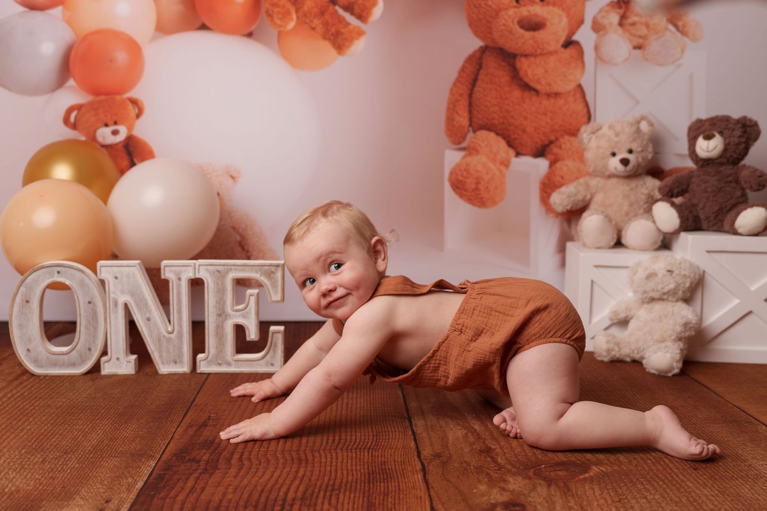 Orange-toned first birthday scene with balloon arch and teddy bear decorations.