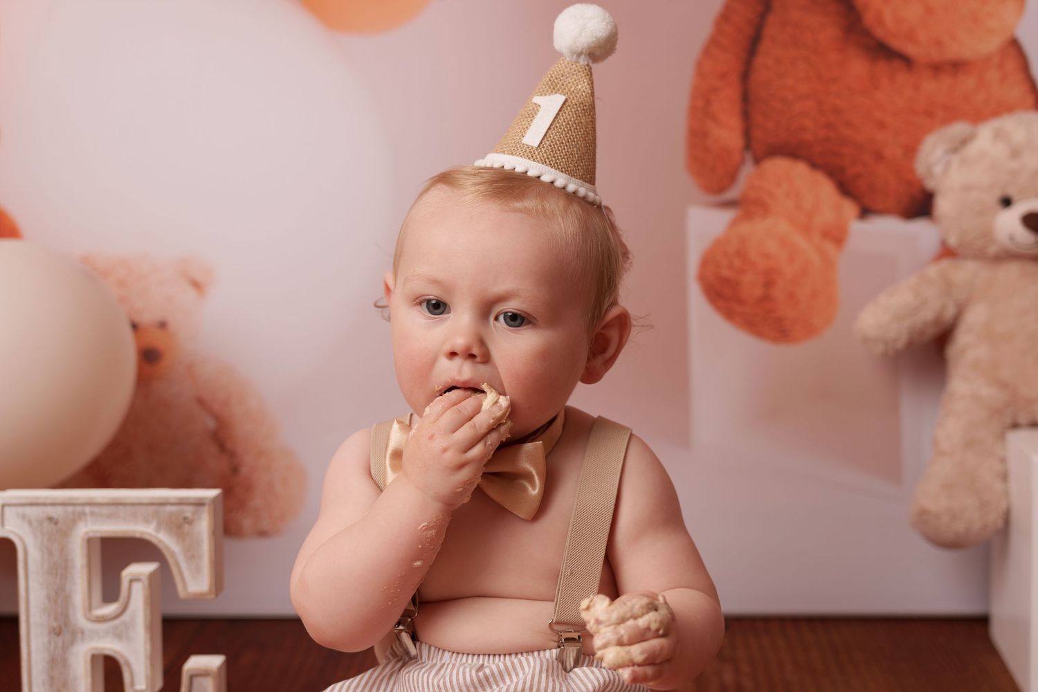 Baby in party hat explores cake during first birthday photoshoot with teddy bears and balloons in background.