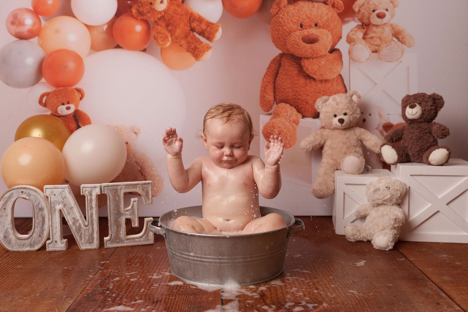 Baby sits in metal tub surrounded by orange and white balloons and teddy bears for first birthday shoot.
