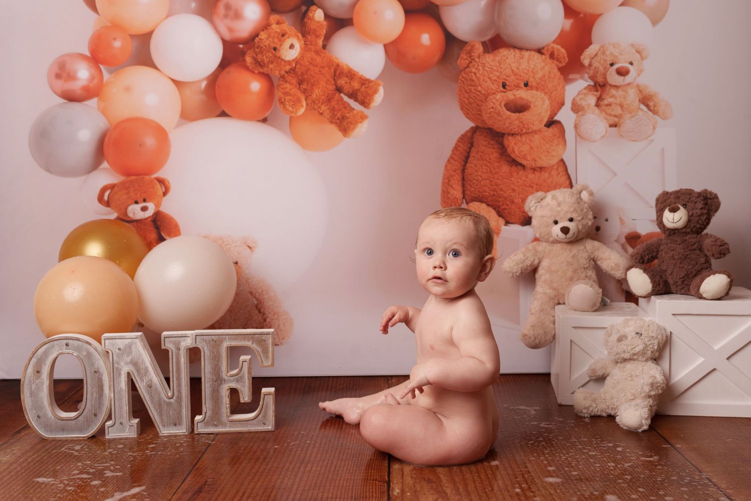 Baby sitting on floor with ONE sign, balloons, and teddy bears for first birthday photoshoot.