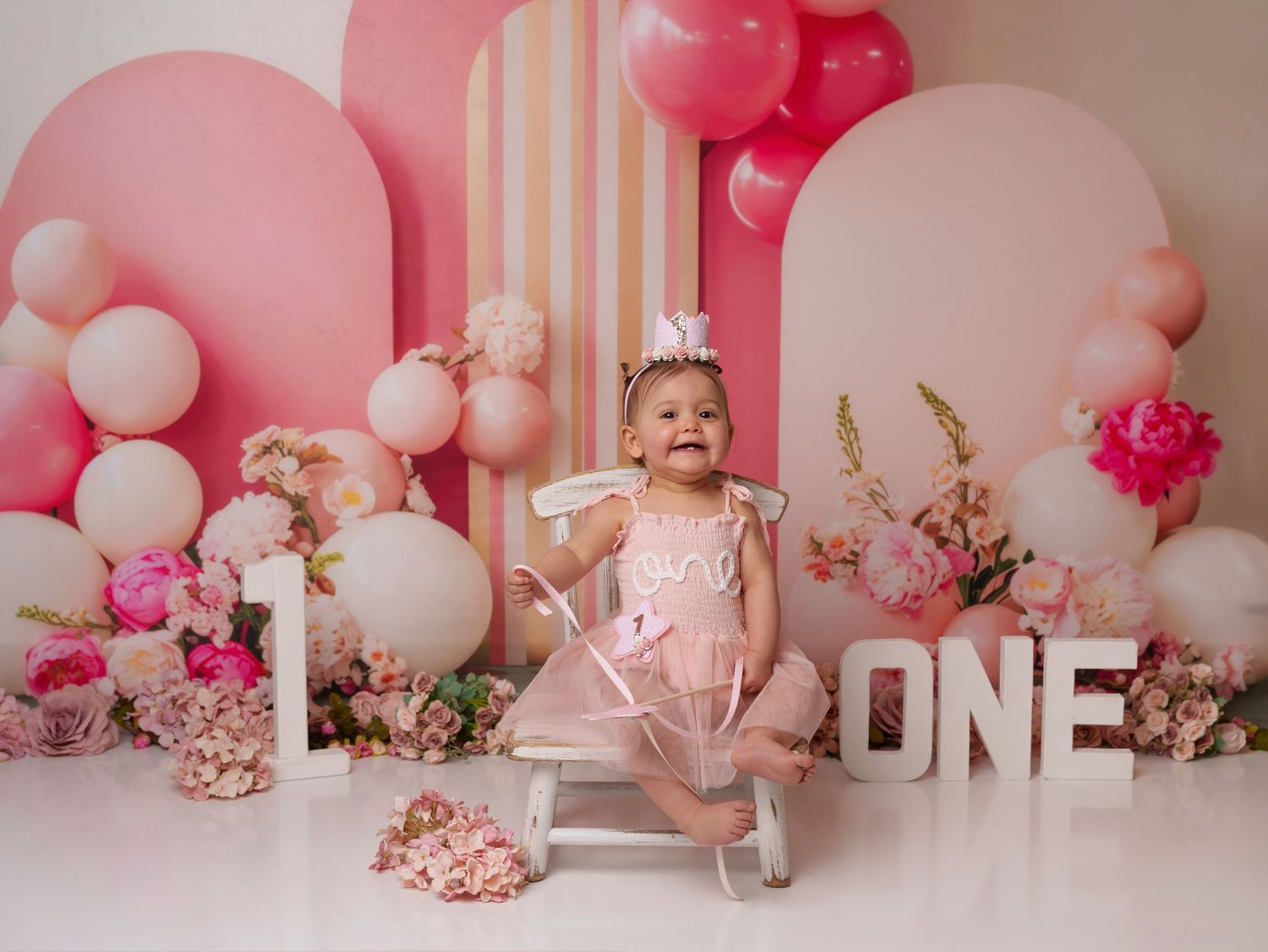 Pink birthday celebration setup with 'ONE' letters, balloons, flowers and decorative backdrop for first birthday photoshoot.