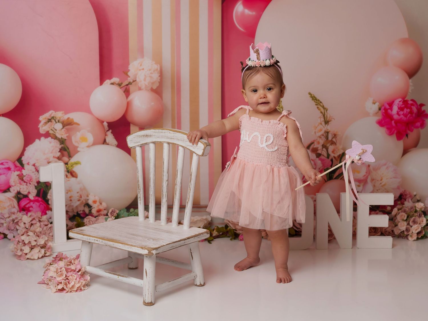 Pink and white first birthday photoshoot setup with balloons, flowers, and a vintage white chair against pastel backdrop.