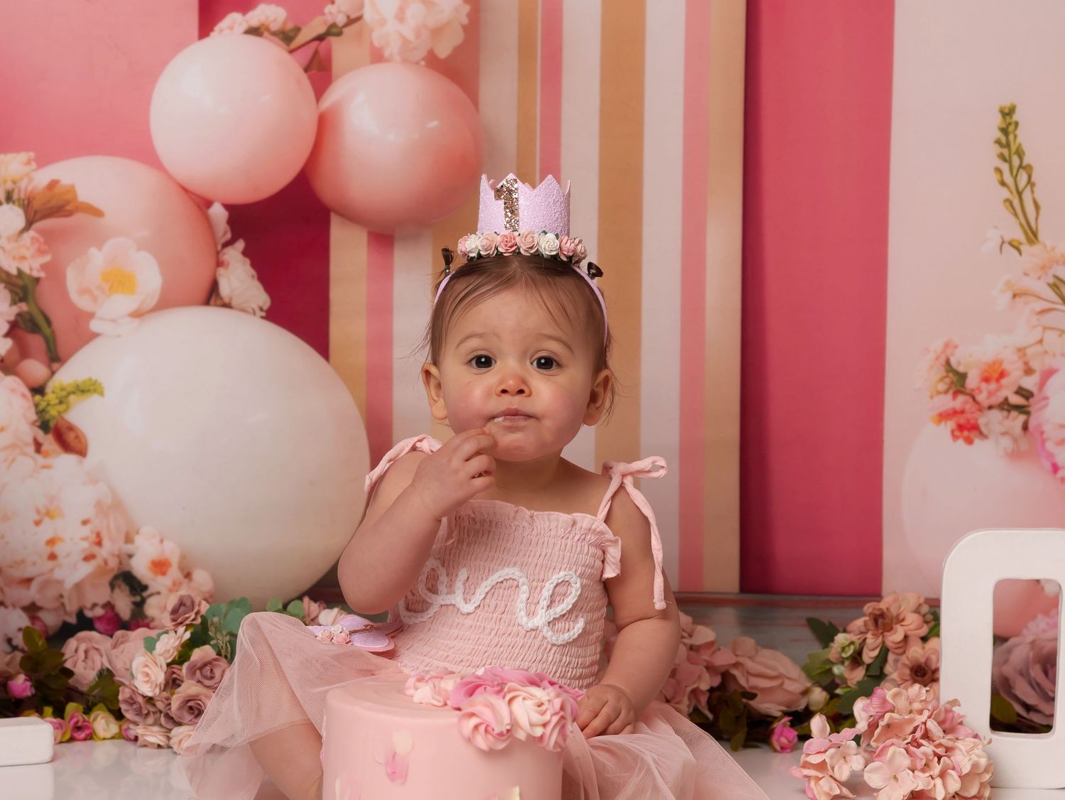 Pretty pink first birthday party setup with balloons, tulle dress, and floral decorations against a striped backdrop.
