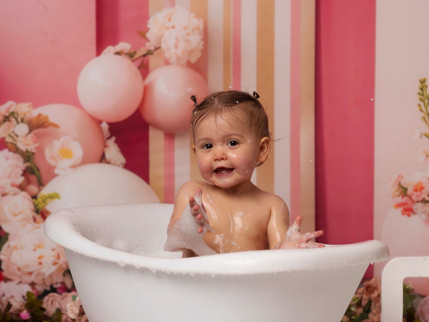 Baby splashing in white bathtub during pink-themed photoshoot with balloons and floral decorations.
