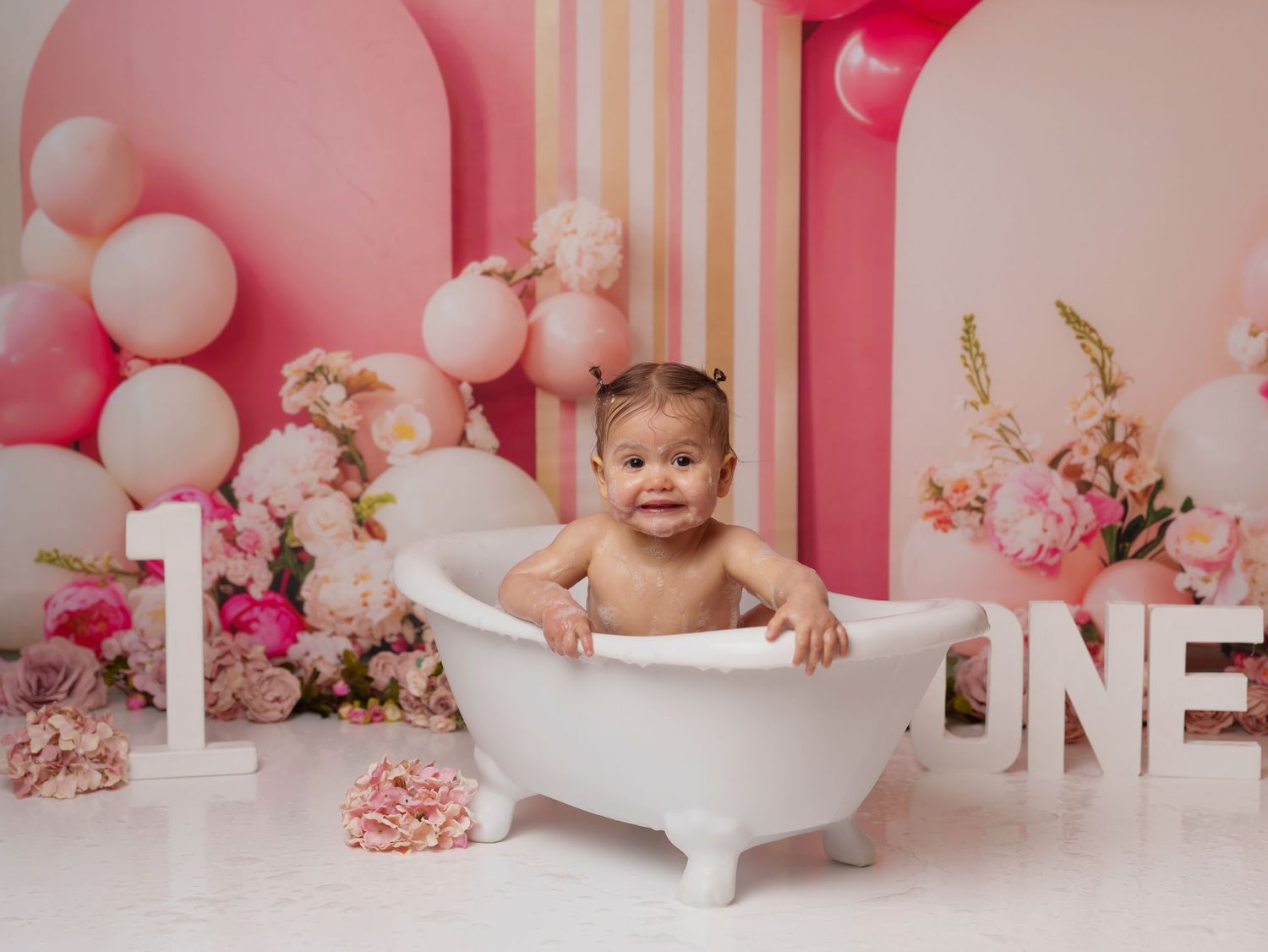 A baby takes a bath in a vintage tub surrounded by pink balloons, flowers and large white ONE letters for first birthday setup.