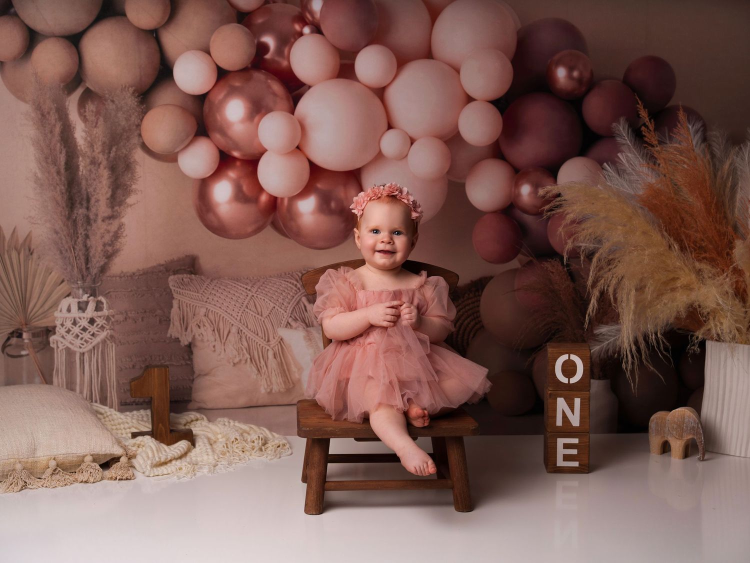 Pink and rose gold balloon arch decorates a dreamy first birthday setup with wooden chair and tulle details.