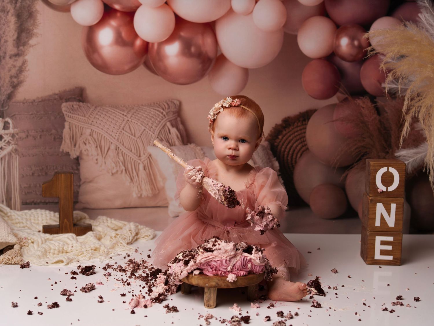 Balloon arch decorates a pink first birthday cake smash setup with wooden blocks spelling ONE and scattered cake crumbs.