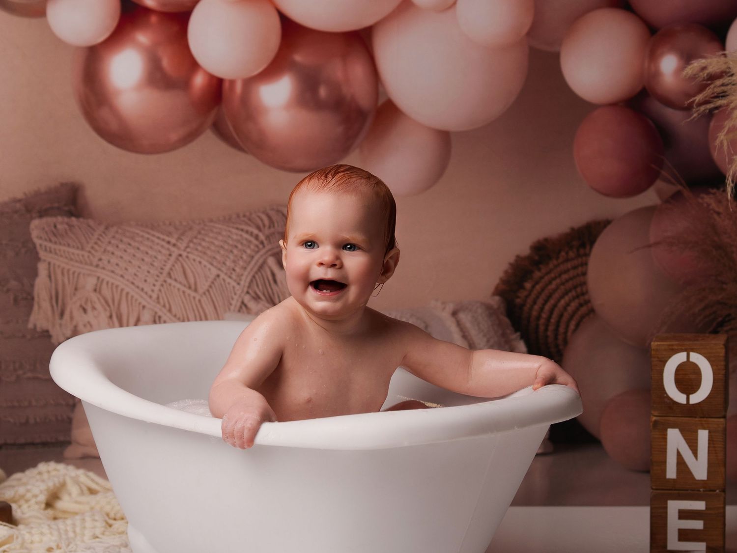 Happy baby sits in white bathtub with rose gold balloon decorations and pink backdrop for first birthday photoshoot.