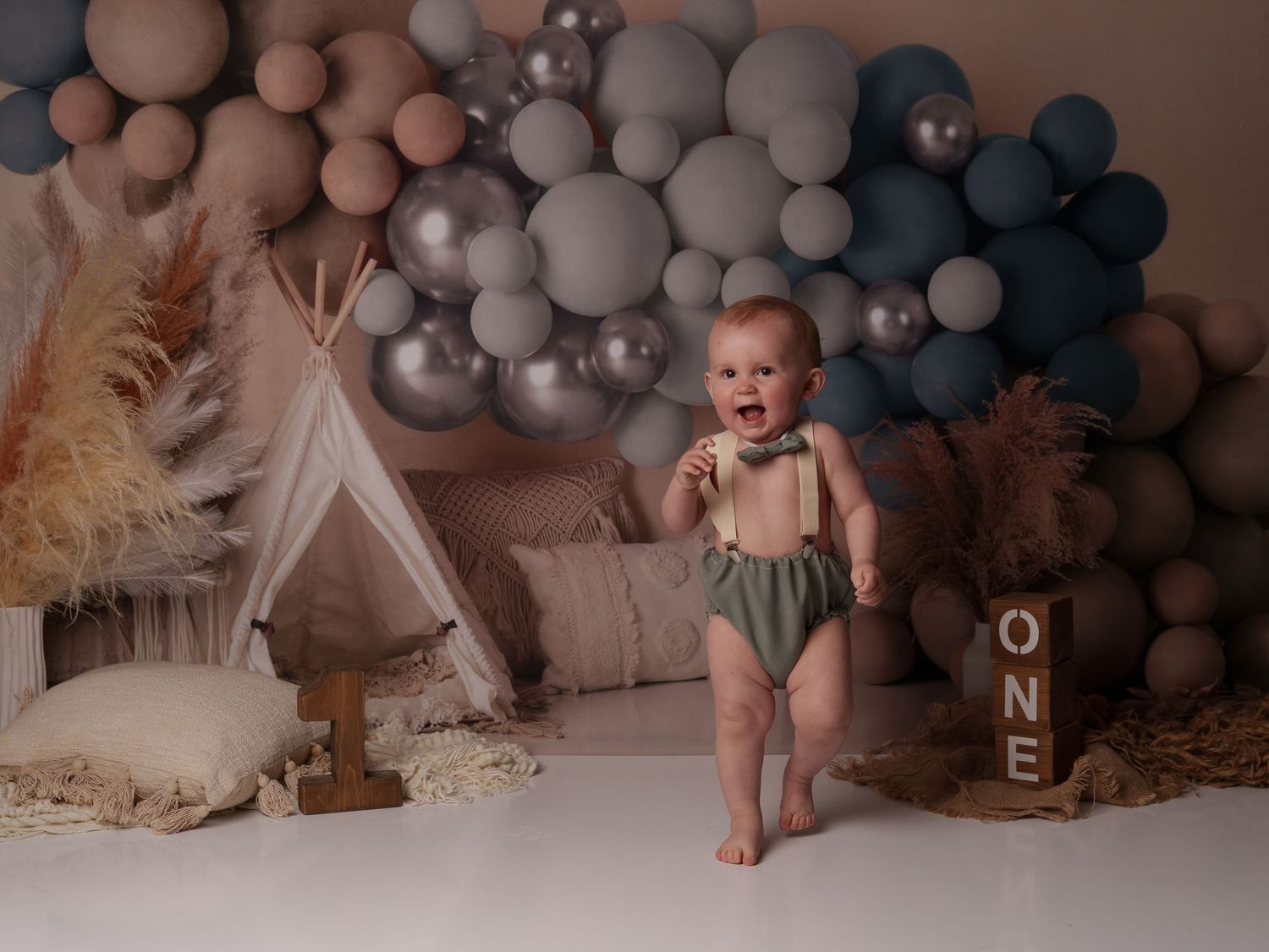 Baby's first birthday photoshoot with gray and blue balloon arch, teepee, and pampas grass decorations.