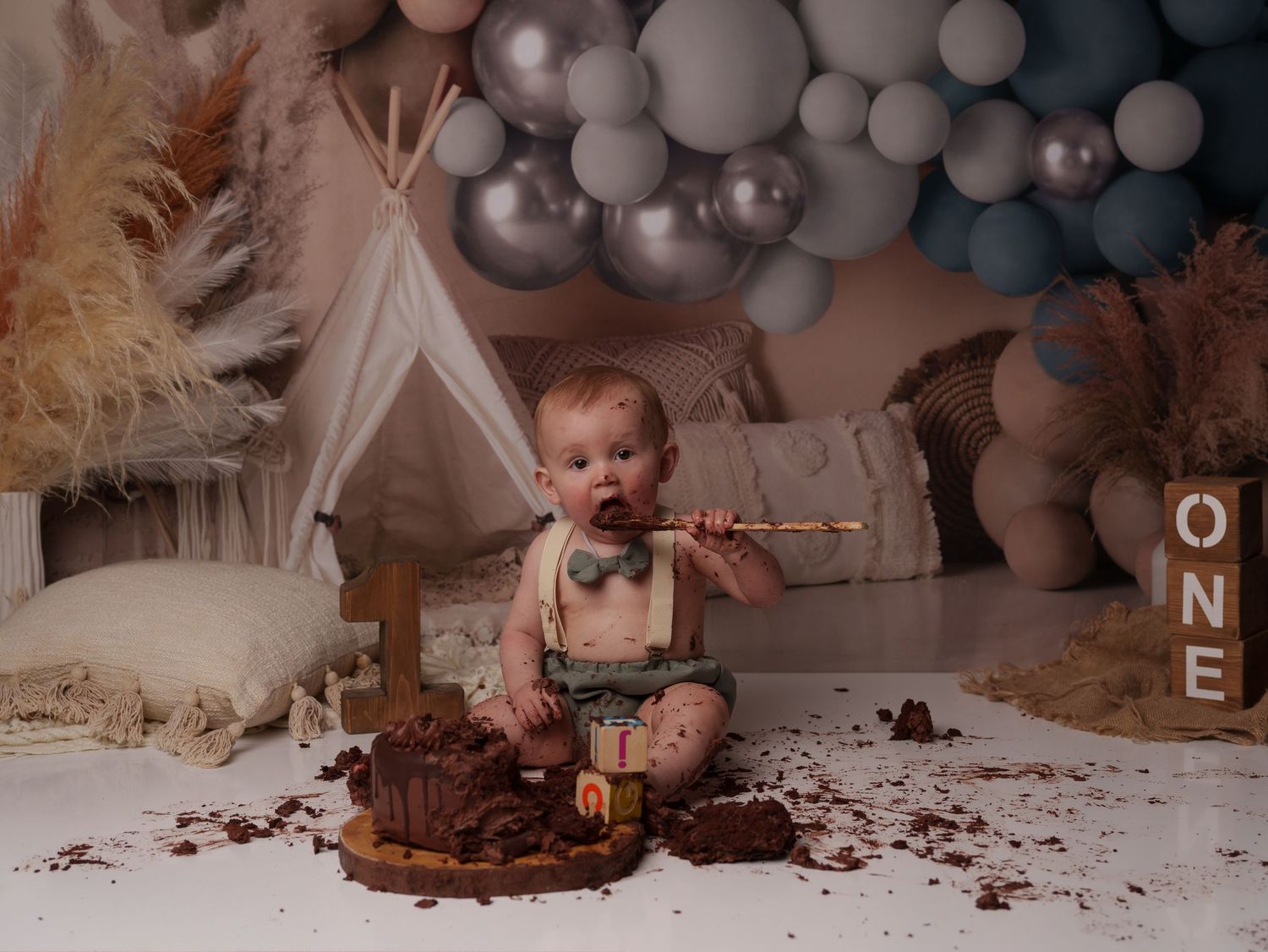 A cake smash celebration setup with white teepee, balloon arch, and scattered chocolate cake against a neutral backdrop.