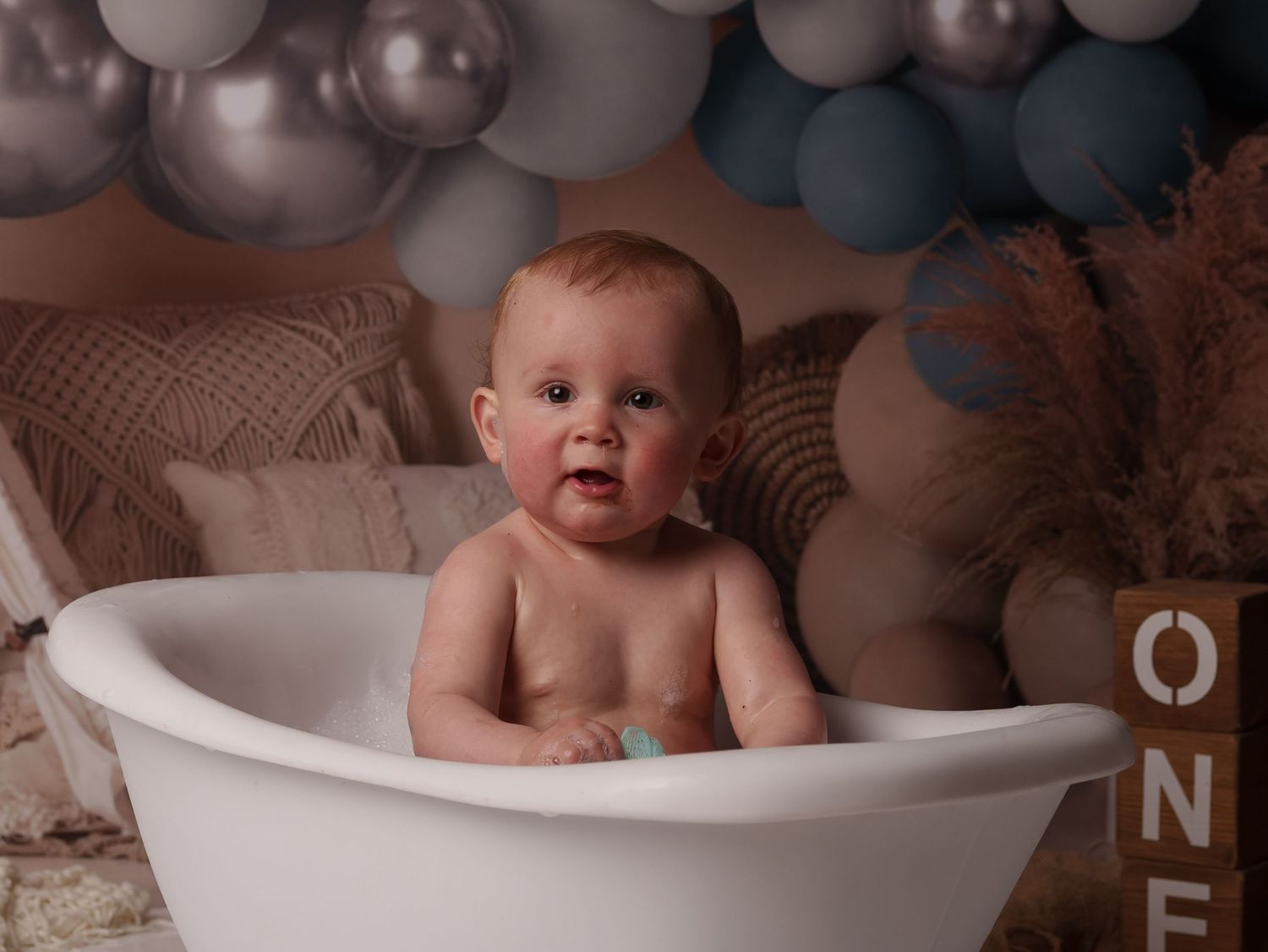 Baby sitting in white bathtub with gray and blue balloons in background during professional photoshoot.