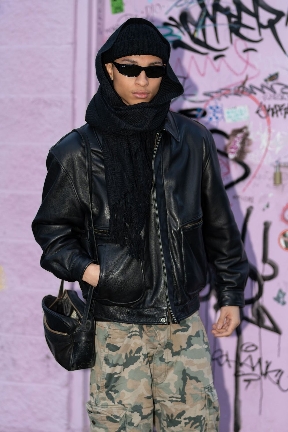 Person wearing leather jacket, black scarf, sunglasses and camo pants poses against graffiti wall with pink backdrop.
