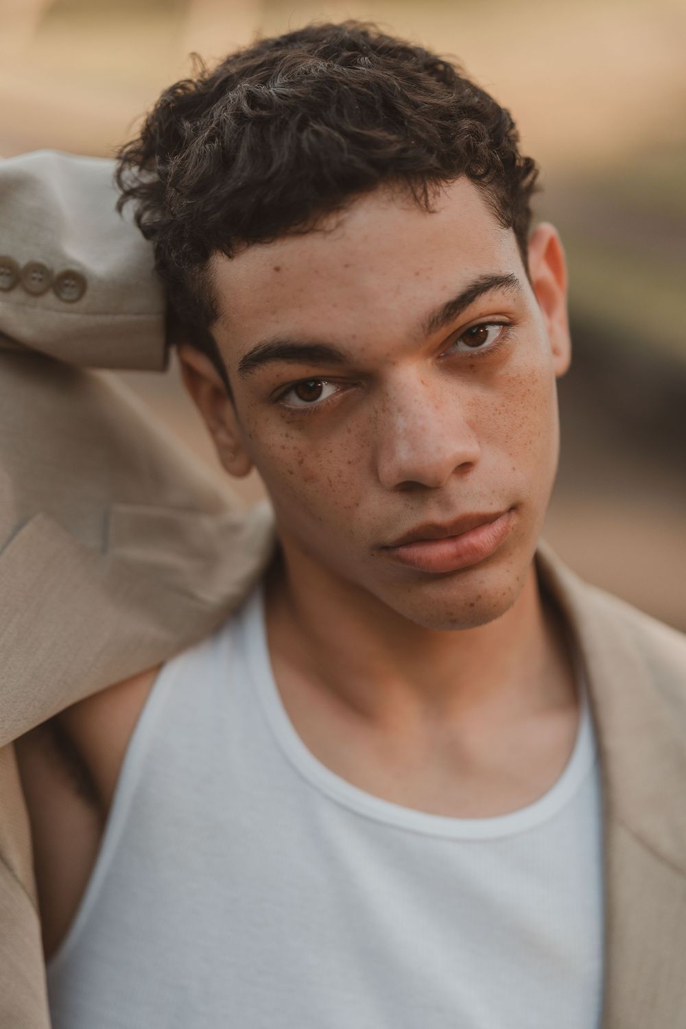Close up portrait showing curly dark hair and freckles wearing a white tank top and beige blazer in warm lighting.
