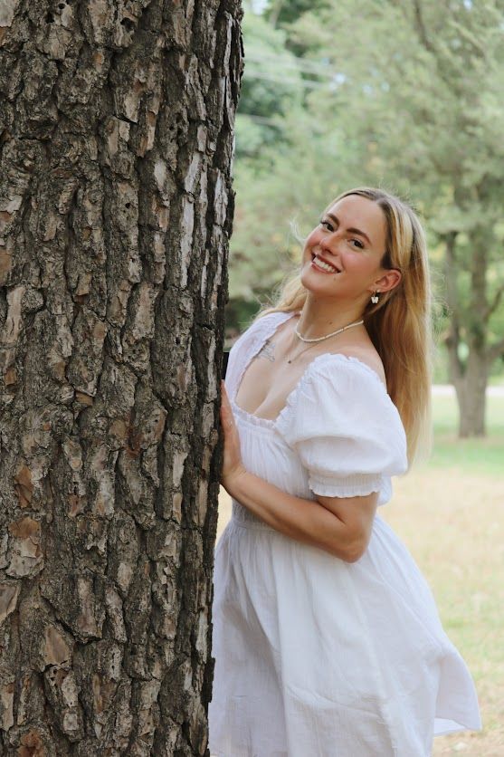 Smiling blonde woman in white off-shoulder dress leaning against a tree trunk in a lush green park.