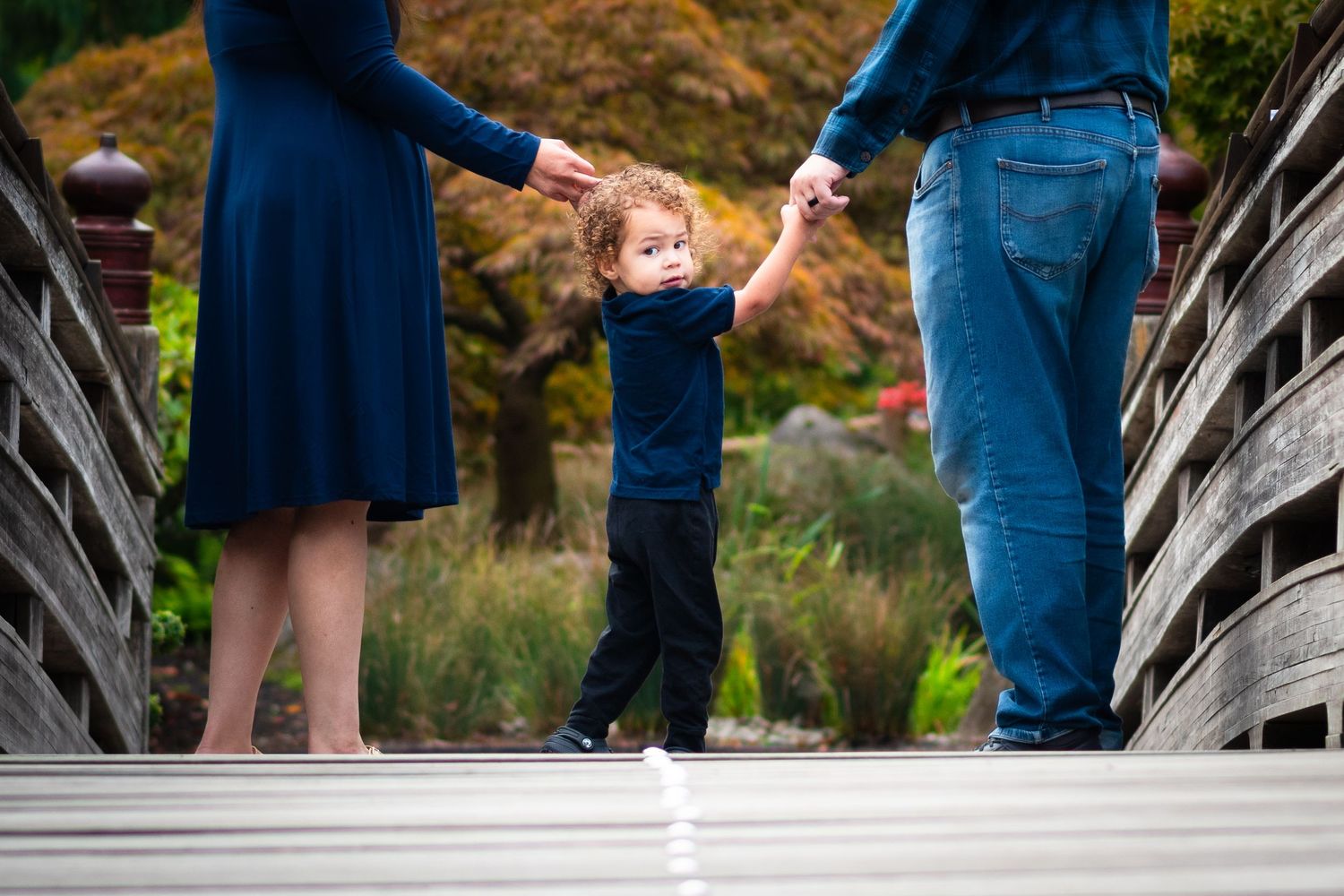 Parents holding hands with child walking on wooden bridge in nature setting.