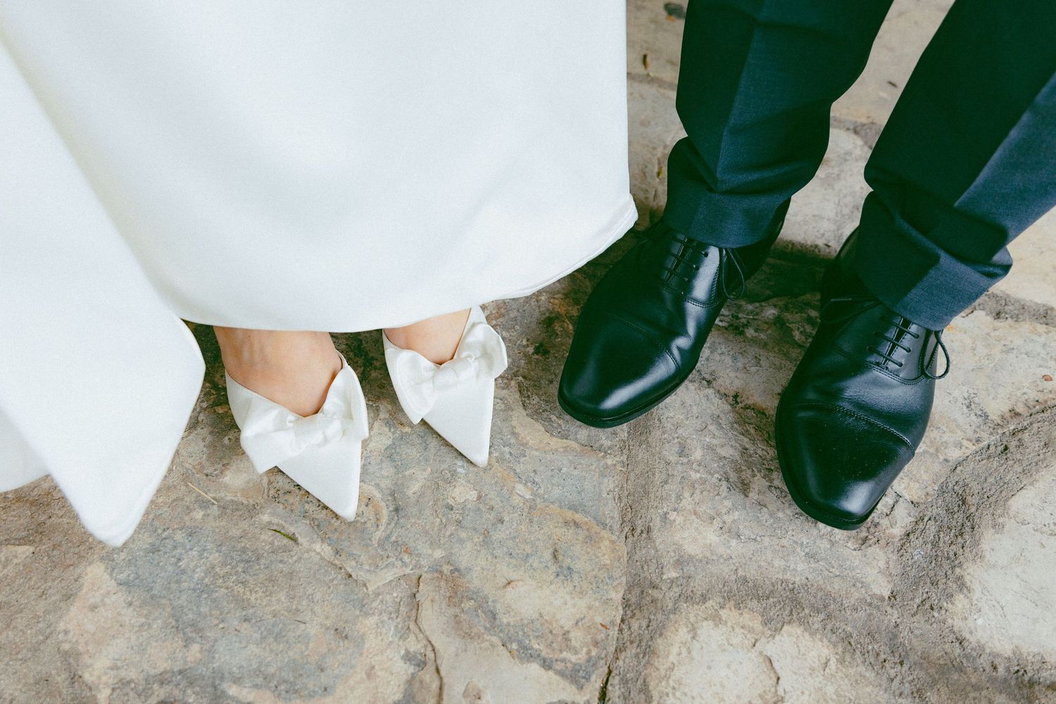 Close-up view of formal black dress shoes and white wedding dress hem against a textured stone floor.
