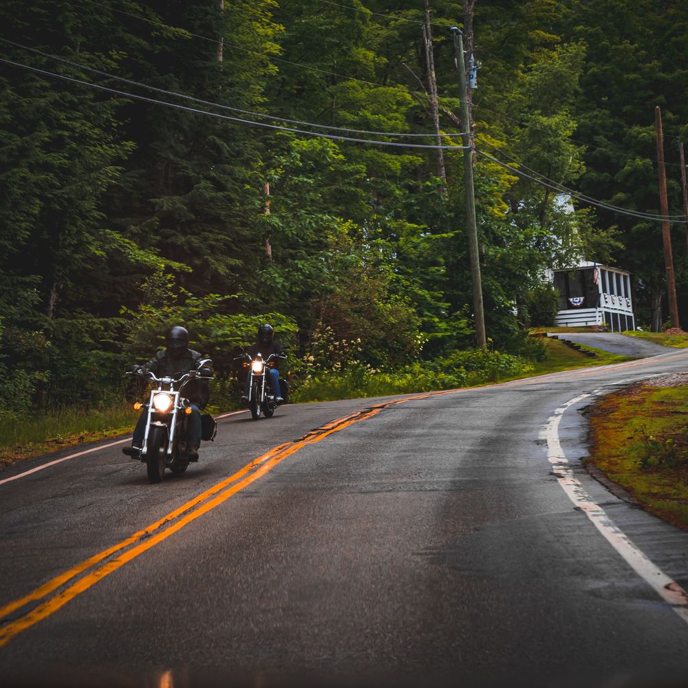 Motorcycles riding in formation along a winding forest road on a cloudy day.