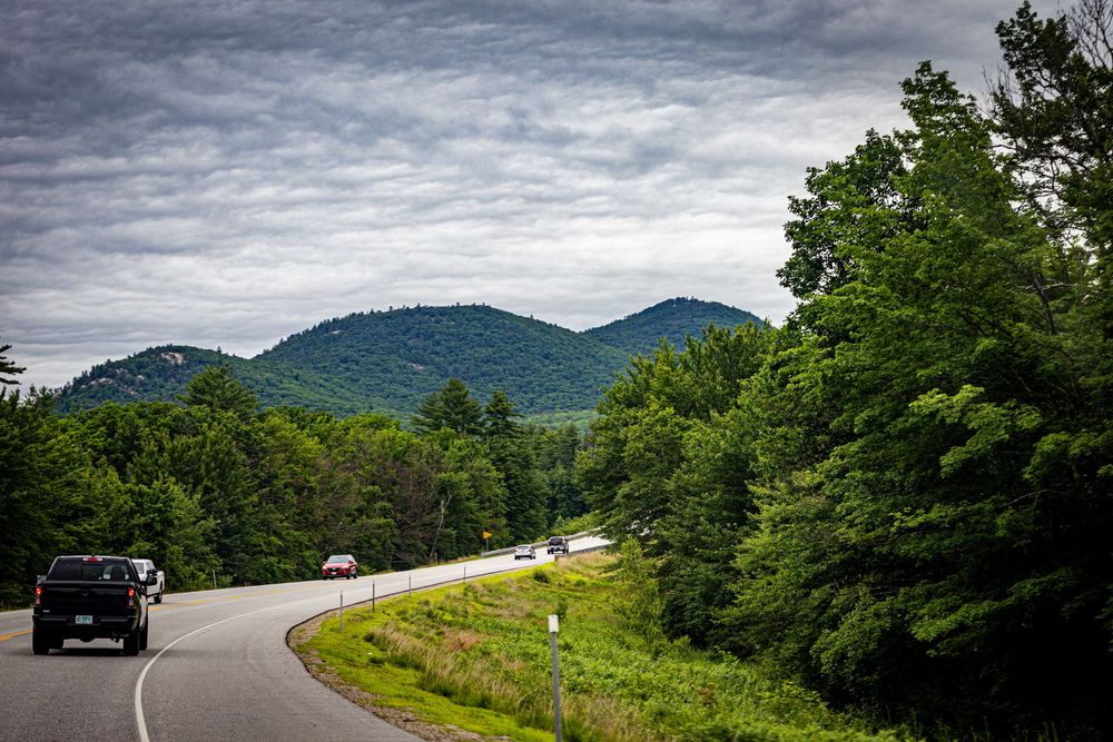 A winding mountain road curves through lush green forests and hills under a dramatic cloudy sky.