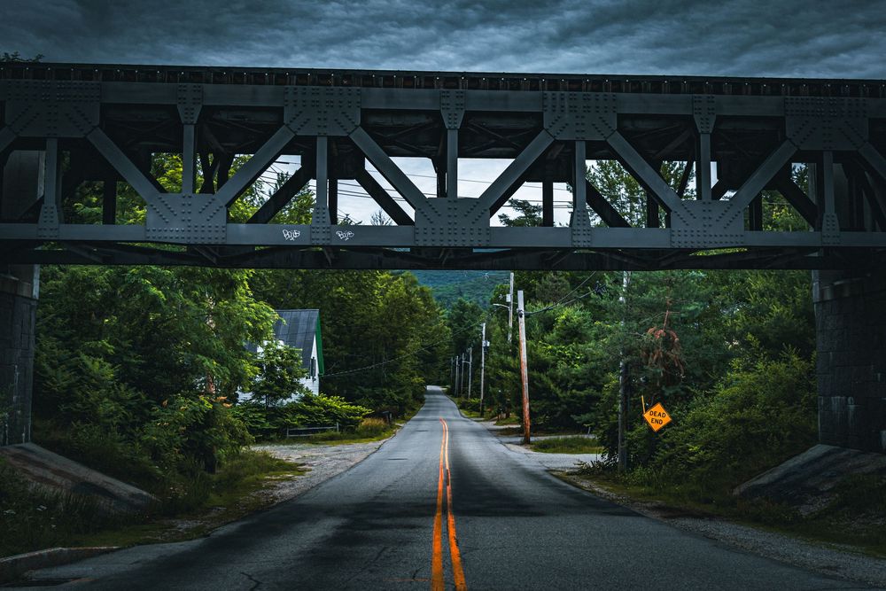 Dark railroad bridge spans over a rural two-lane road with yellow dividing lines, surrounded by green forest.