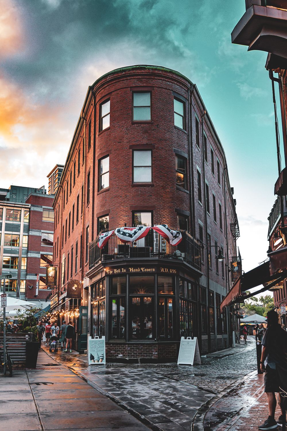 Historic red brick building with curved corner facade and British flags in Boston's North End during sunset with moody sky.