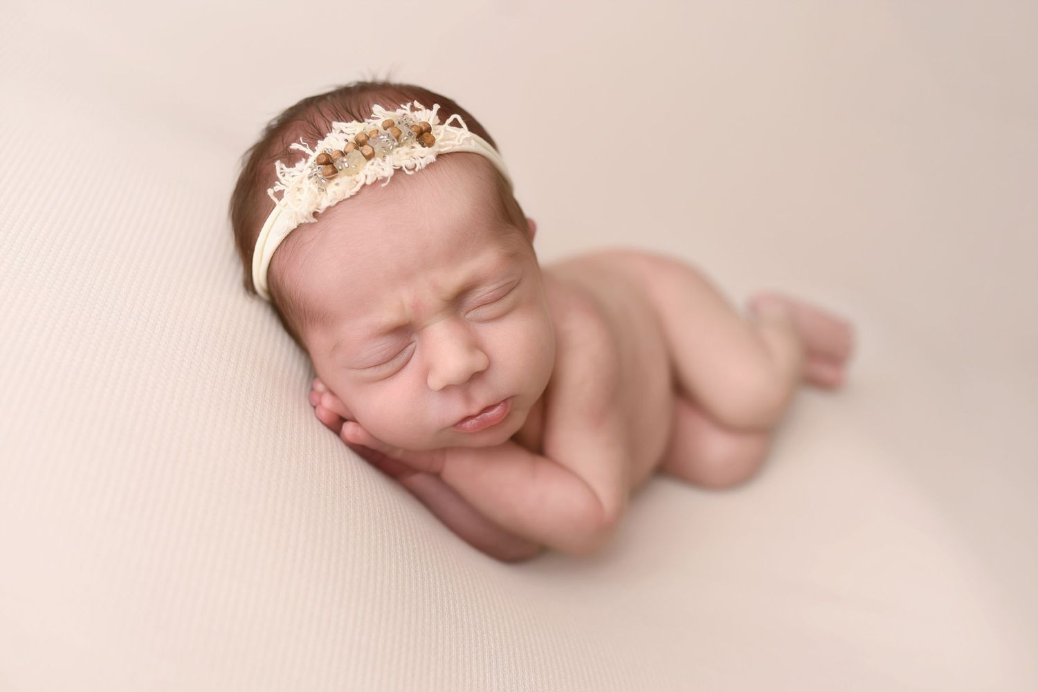 Peaceful sleeping newborn girl wearing delicate crown headband on soft beige backdrop.