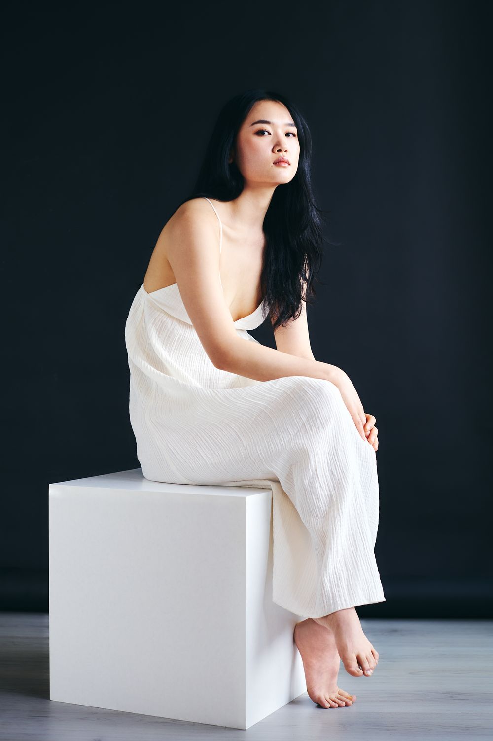 Artistic portrait series showing figure in white fabric seated on minimalist white cube against dark background.
