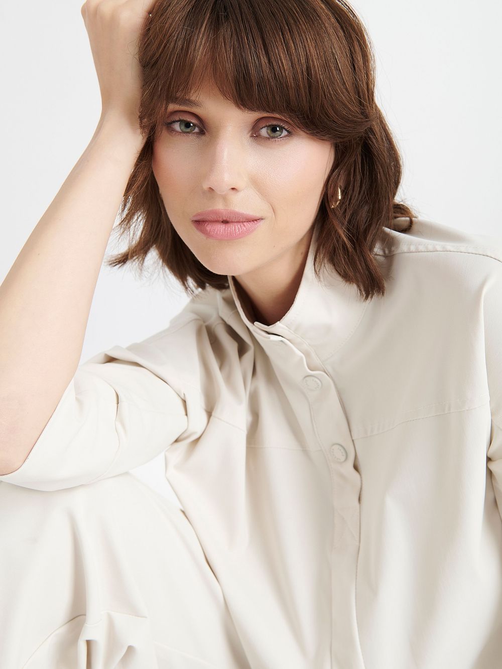 Model wearing a cream colored button-up blouse with long sleeves poses against white background with short brown hair.