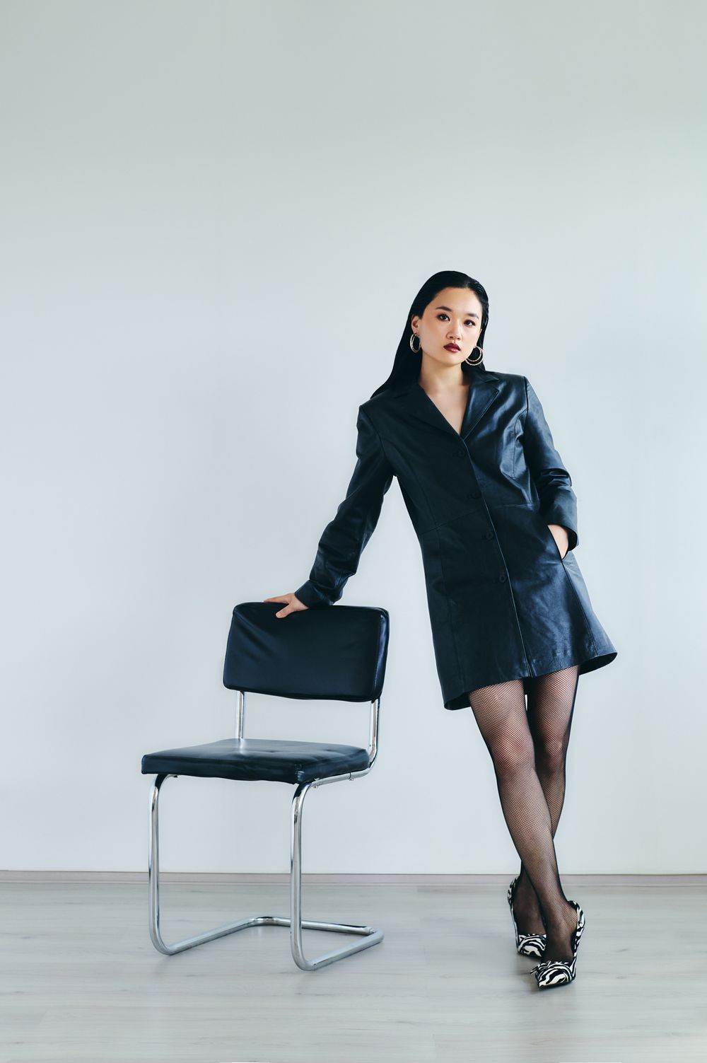 A figure in a black blazer and stockings poses artistically with a modern chrome chair against a white backdrop.