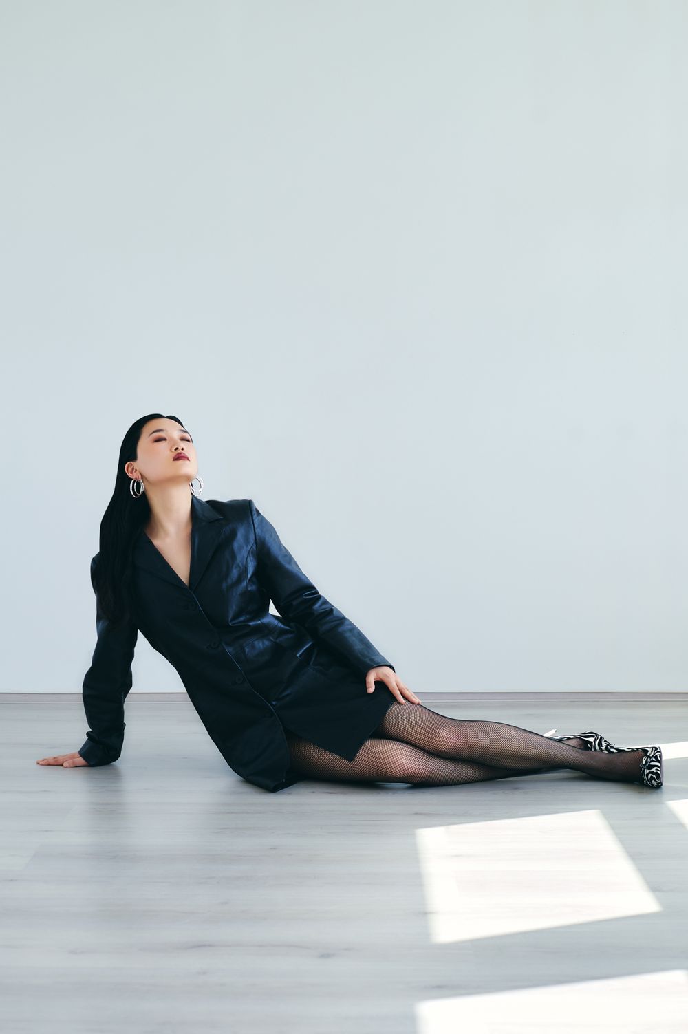 Fashion model in black blazer reclines on white floor in artistic pose with dramatic lighting.
