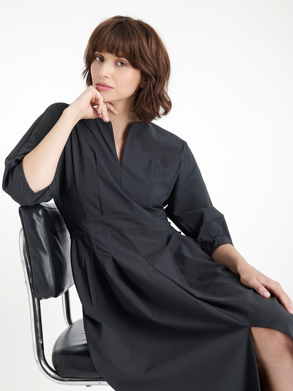 A person in a black kimono-style dress poses thoughtfully on a white chair against a light background.