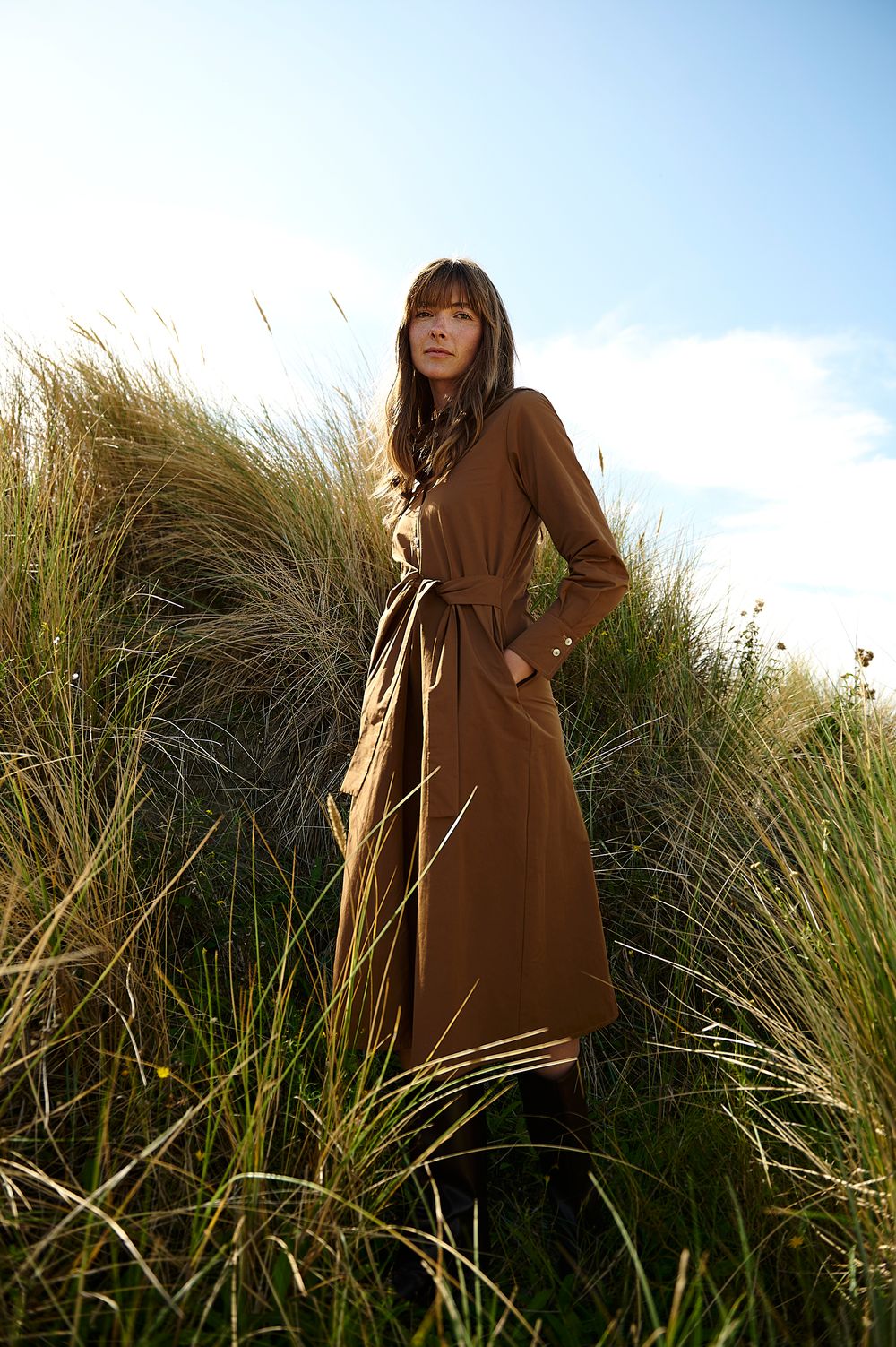 A person in a brown dress stands among tall wild grass against a blue sky background.