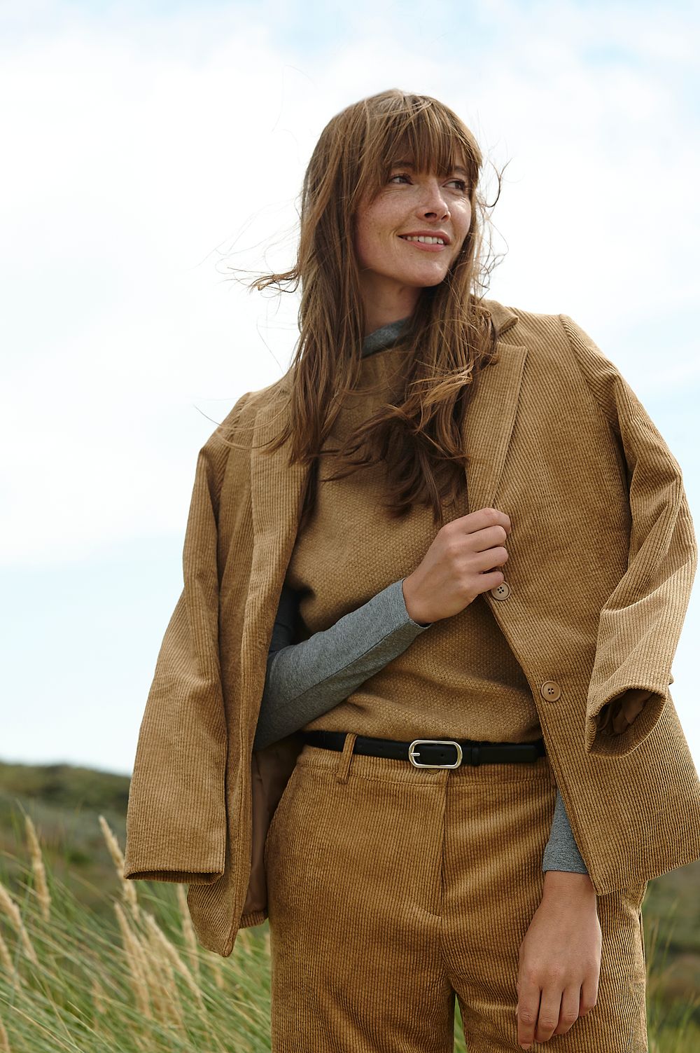 A person wearing a brown corduroy suit and gray sweater poses playfully in a grassy outdoor setting.