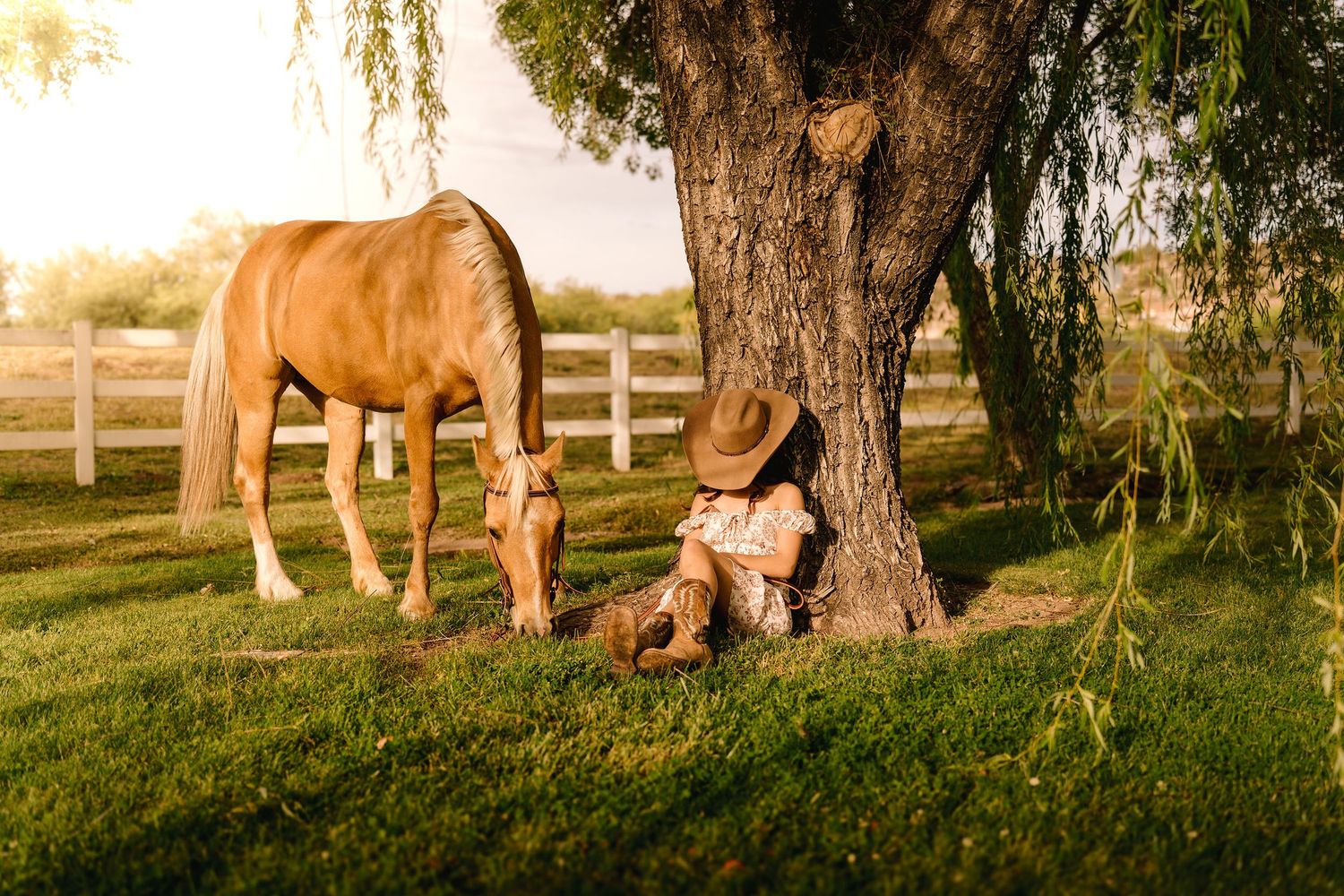 Child in cowboy hat resting against tree beside grazing brown horse on sunny green farm with white fence.