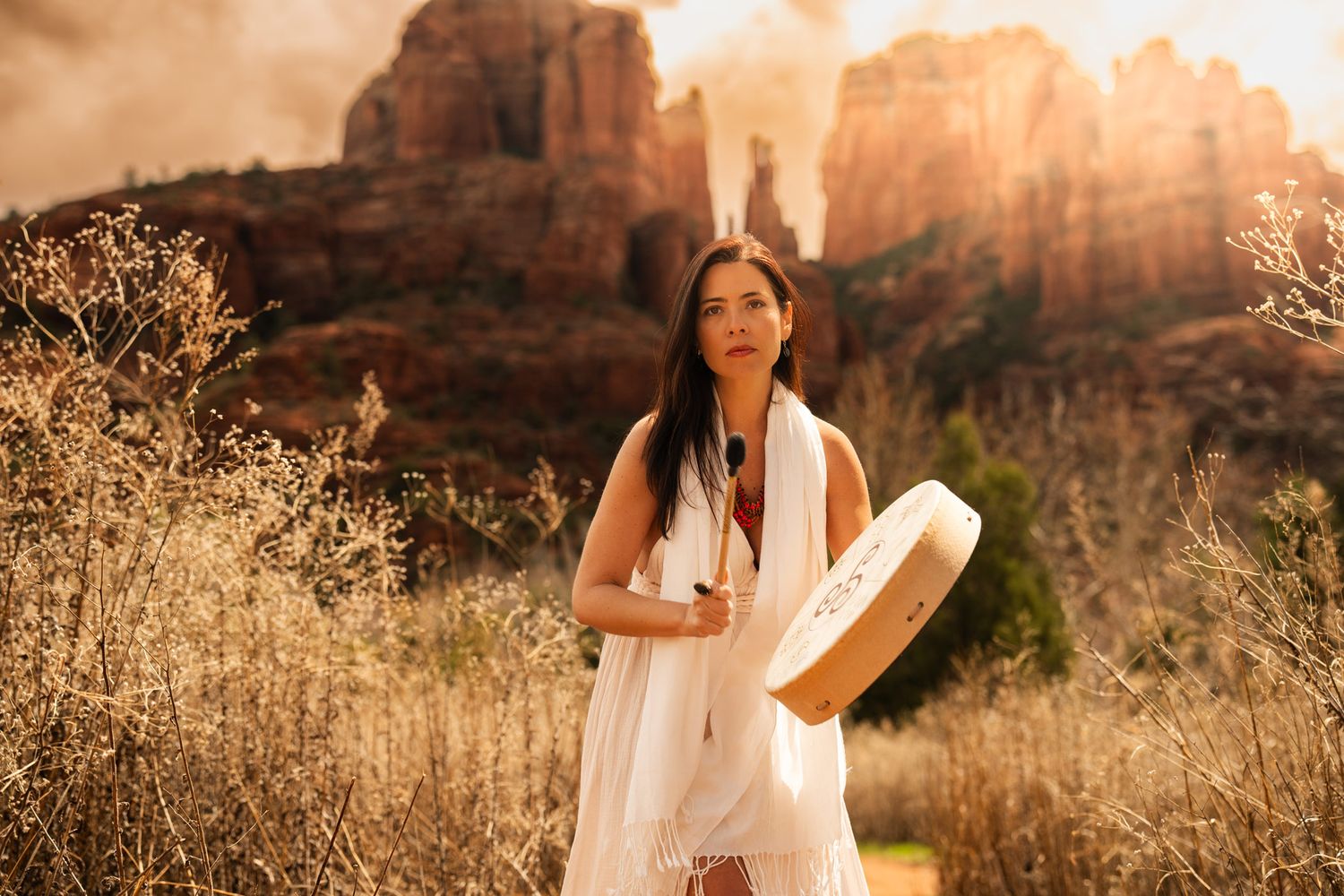 Woman in white dress holds a drum against Sedona desert landscape at golden hour.