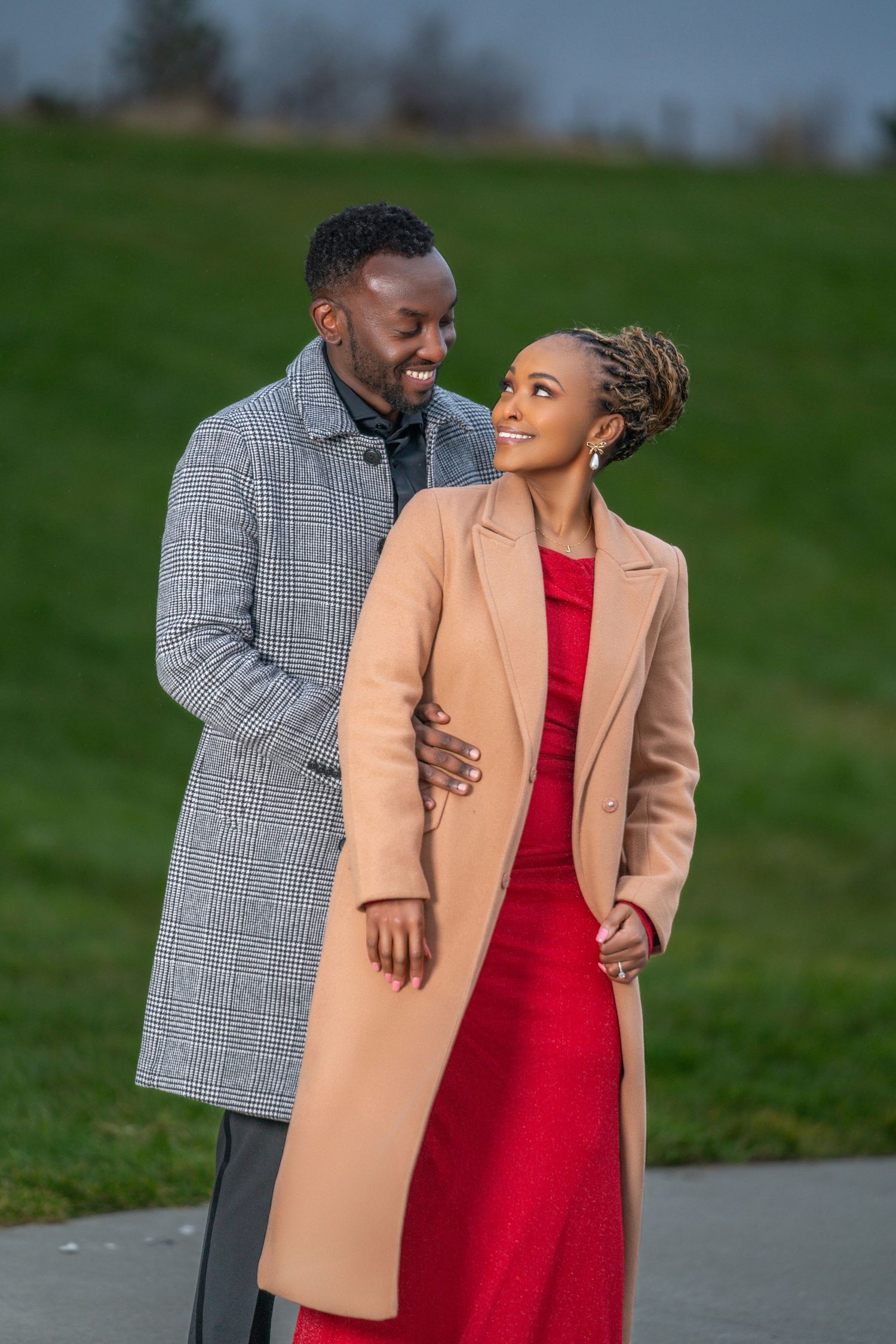 A couple in winter coats walks together outdoors, with one wearing a red dress and tan coat, the other in gray plaid.