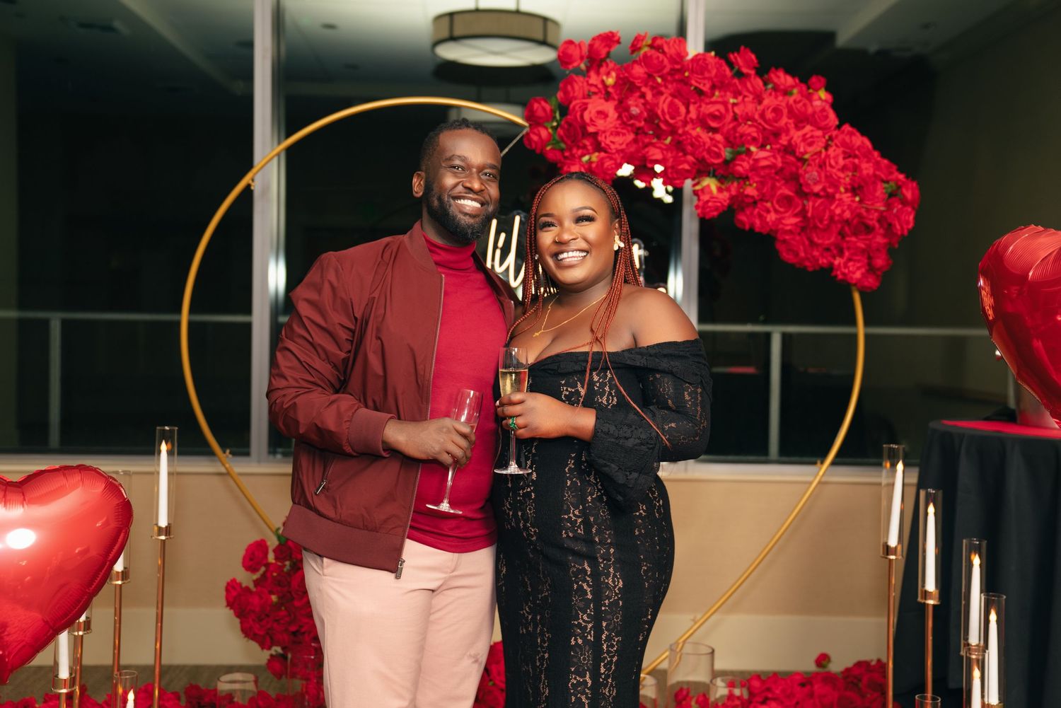 Couple poses in front of a golden arch and red floral arrangement at an elegant Valentine's Day celebration.