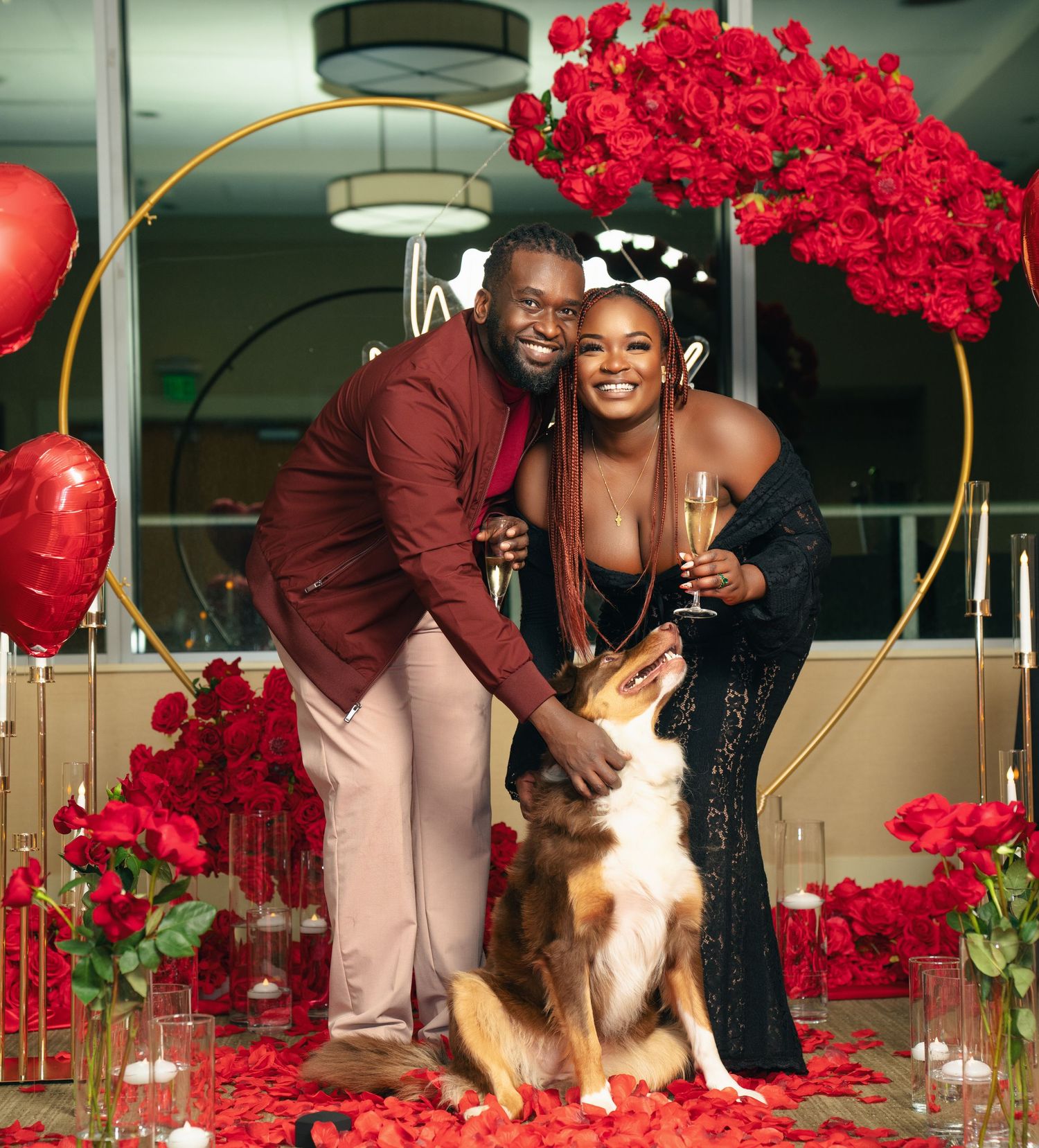 A couple poses in front of a circular gold arch decorated with red roses and balloons, with their German Shepherd dog.